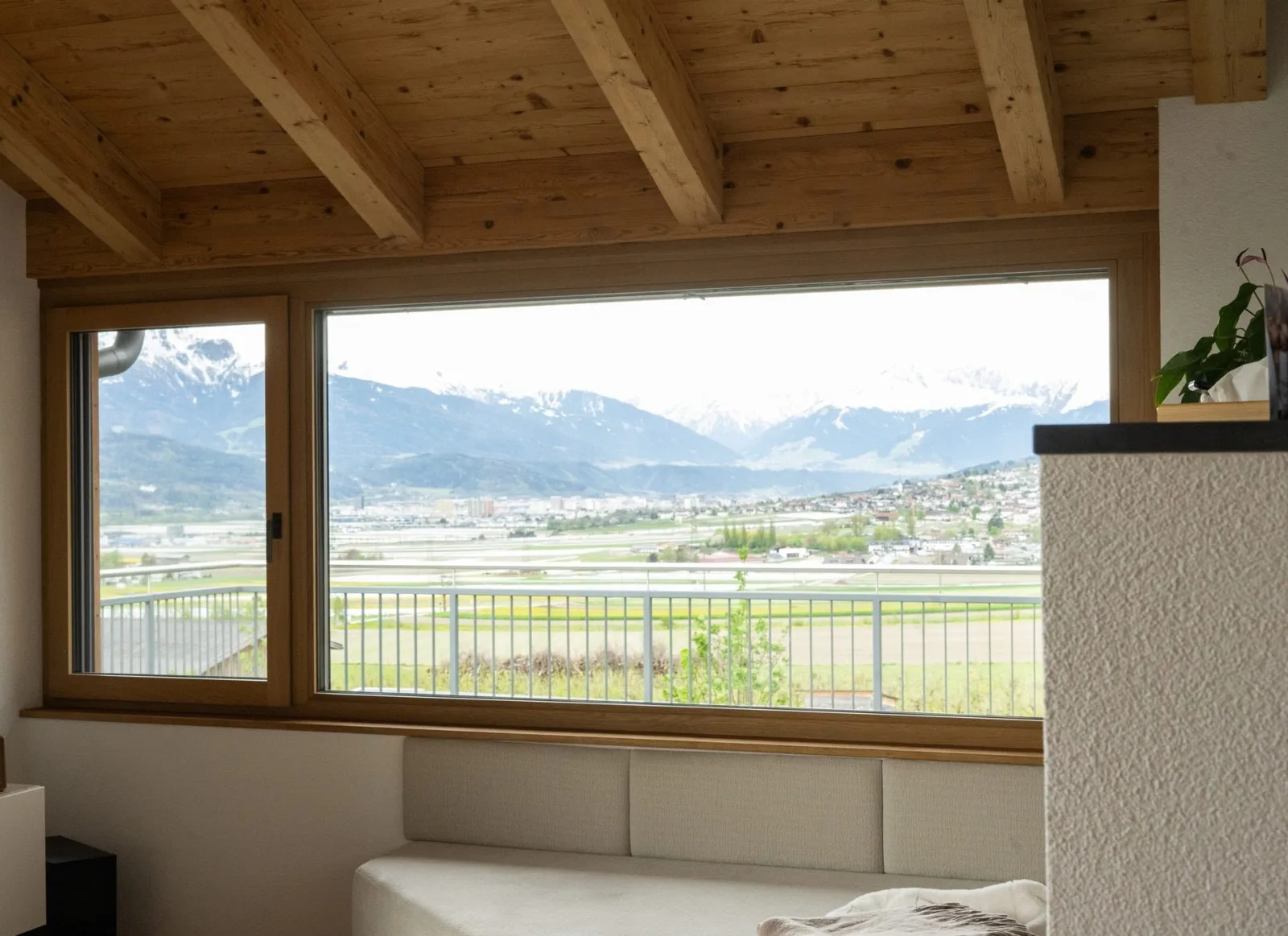 Großes Holzfenster mit Blick über das Tal auf die Berge, davor eine Sitzbank